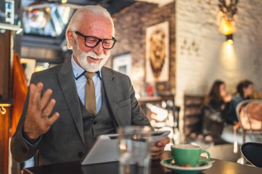 Bir üst düzey Kafkas erkek iş adamı dijital tablet kullanıyor kafede oturup görüntülü konuşma bağlantısı kuruyor ve iletişim kıyafeti fotokopi odası giyiyor.