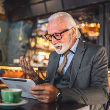 Bir üst düzey Kafkas erkek iş adamı dijital tablet kullanıyor kafede oturup görüntülü konuşma bağlantısı kuruyor ve iletişim kıyafeti fotokopi odası giyiyor.