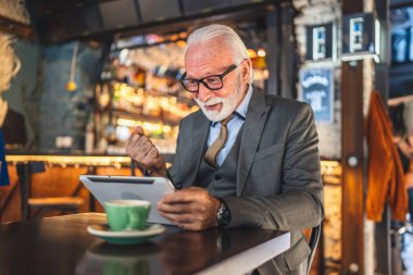Bir üst düzey Kafkas erkek iş adamı dijital tablet kullanıyor kafede oturup görüntülü konuşma bağlantısı kuruyor ve iletişim kıyafeti fotokopi odası giyiyor.