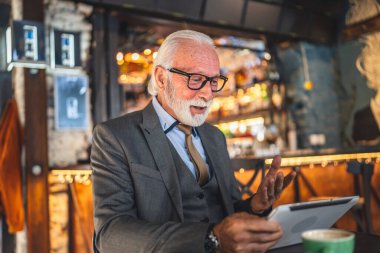 Bir üst düzey Kafkas erkek iş adamı dijital tablet kullanıyor kafede oturup görüntülü konuşma bağlantısı kuruyor ve iletişim kıyafeti fotokopi odası giyiyor.