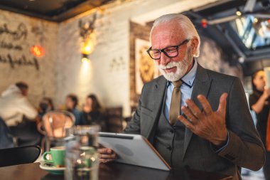 Bir üst düzey Kafkas erkek iş adamı dijital tablet kullanıyor kafede oturup görüntülü konuşma bağlantısı kuruyor ve iletişim kıyafeti fotokopi odası giyiyor.