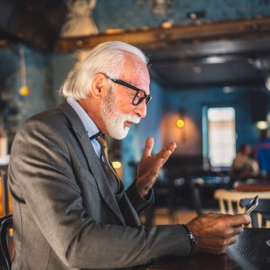 Bir üst düzey Kafkas erkek iş adamı dijital tablet kullanıyor kafede oturup görüntülü konuşma bağlantısı kuruyor ve iletişim kıyafeti fotokopi odası giyiyor.