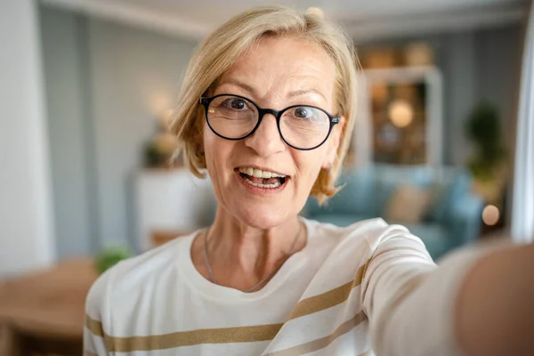 一个成熟的金发碧眼的女人的画像 带着眼镜在家里开心地笑着看着相机自信地穿着毛衣在明亮的房间里用户生成的内容自画像自我复制的空间 — 图库照片