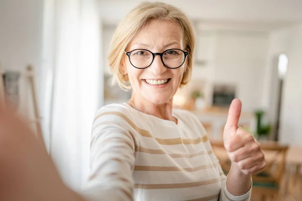 1成熟したブロンドCaucasian女性の肖像画とともに眼鏡とともに自宅で幸せな笑顔見ますカメラに自信を持って着用セーターで明るい部屋ユーザー生成コンテンツ自画撮りUgcコピースペース — ストック写真