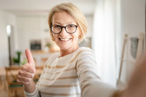 1成熟したブロンドCaucasian女性の肖像画とともに眼鏡とともに自宅で幸せな笑顔見ますカメラに自信を持って着用セーターで明るい部屋ユーザー生成コンテンツ自画撮りUgcコピースペース — ストック写真