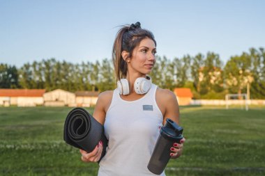 one woman adult caucasian female athlete standing on stadium on running track with supplement shaker and yoga mat with headphones ready for training outdoor in summer evening real people copy space