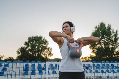 Bir yetişkin beyaz kadın sporcu yaz akşamları stadyumda kettlebell girya ağırlığı ile antrenman yapıyor. Kulaklıklarıyla mutlu esmer sağlık ve zindelik konsepti fotokopisini çekiyor.