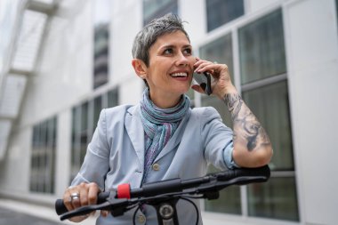 Yetişkin bir yaşlı beyaz kadın dışarıda duruyor. Cep telefonu kullanıyor. Gündüz güvenli modern alternatif ulaşım ve iletişim alanı kopyalama yönteminde elektrikli scooterla konuşuyor.