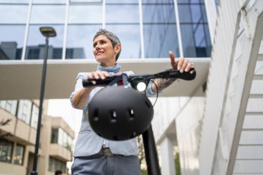 Bir kadın olgun, yaşlı, beyaz, açık havada elektrikli scooterlı bir kadın. Gündüz kendine güvenen bir iş kadını modern alternatif ulaşım yöntemini kullanıyor. Gerçek insanlar uzayı kopyalıyor.
