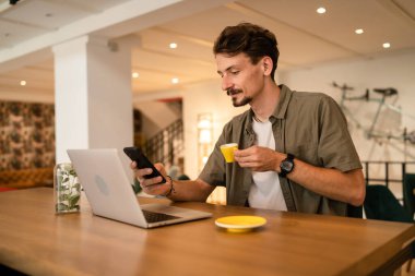 one caucasian man young adult millennial male using mobile phone stand at table in cafe at the table alone happy smile confident having online chat using internet for text messages while work remote