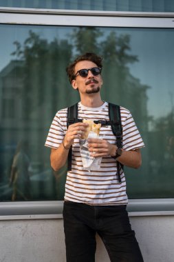 Genç, yetişkin, beyaz bir erkek, güneşli bir günde şehirde duruyor ve sandviç yiyor. Fast food konsepti. Şehir hayatı.