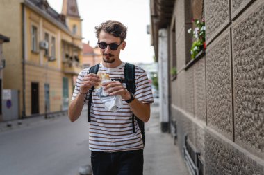 Gündüz vakti şehirde genç, yetişkin, beyaz bir erkek sandviç yiyor. Fast food konsepti. Şehir hayatı.