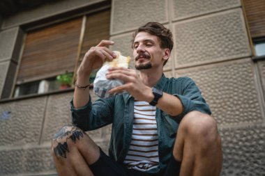 Genç, yetişkin, beyaz bir erkek, güneşli bir günde şehirde duruyor ve sandviç yiyor. Fast food konsepti. Şehir hayatı.