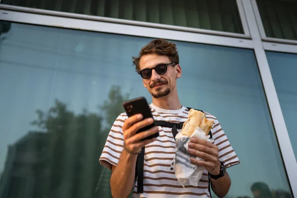 晴れた日のスタンドで市内の1人の若い大人の現代的な白人男性とサンドイッチを食べますファーストフードのコンセプト都市生活コピースペース観光客が建物の壁の前で食べる本物の人が携帯電話を使用します — ストック写真
