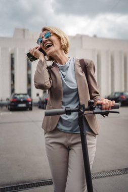 Yetişkin, beyaz, sarışın bir kadın dışarıda elektrikli tekmeyle dikiliyor scooter 'ı itiyor, cep telefonuyla telefon görüşmesi yapıyor şehirde tek başına kendine güveni tam, gerçek insanlar mekanı kopyalıyor.
