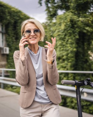 Yetişkin, beyaz, sarışın bir kadın dışarıda elektrikli tekmeyle dikiliyor scooter 'ı itiyor, cep telefonuyla telefon görüşmesi yapıyor şehirde tek başına kendine güveni tam, gerçek insanlar mekanı kopyalıyor.