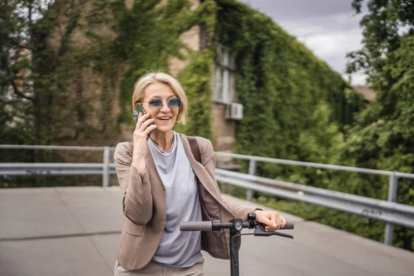 1人の女性成熟したCaucasianブロンド女性スタンド屋外で電気キックプッシュスクーター使用携帯電話スマートフォンで市内の通話を作るために単独で自信を持って現実の人々コピースペース — ストック写真