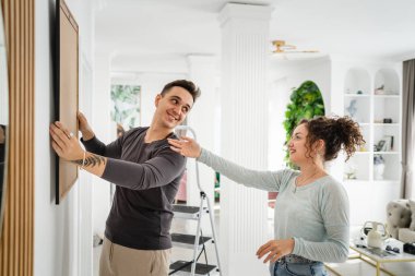 Happy couple caucasian man and woman husband and wife or boyfriend and girlfriend hanging painting on the wall at home in their apartment real people family domestic life concept copy space