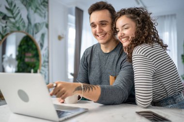 İki yetişkin genç çift karı koca ya da karı koca ya da kız arkadaş mutfakta dizüstü bilgisayarla çevrimiçi içerik alışverişini kontrol ediyorlar ya da video sabah rutini yapıyorlar.