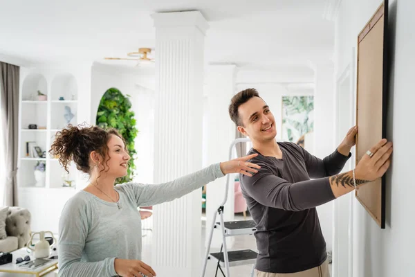 stock image Happy couple caucasian man and woman husband and wife or boyfriend and girlfriend hanging painting on the wall at home in their apartment real people family domestic life concept copy space