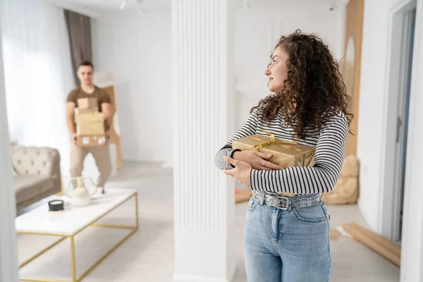 woman young adult caucasian female standing at home with gift box presents holding in hands while her boyfriend or husband man is approaching coming behind real people celebrate surprise concept