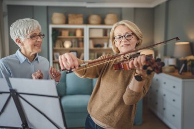 Two women mature female teacher give private violin lesson to her student or friend at home help her to learn to play in room hobby and leisure active senior concept copy space clipart