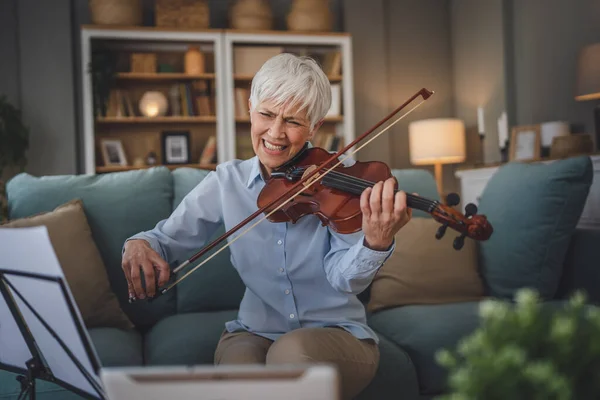 Mature senior caucasian woman learn to play violin practice at home online professor teacher teach or have internet private class on digital tablet while sit on sofa bed at home active senior hobby