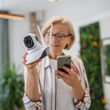 Yetişkin bir beyaz kadın modern güvenlik kamerası ve cep telefonunu elinde tutuyor. Uygulama kopyalama alanı kurmaya ve ayarlamaya çalışıyor.