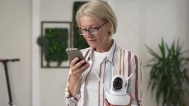 Modern güvenlik kamerası ve cep telefonunu elinde tutan yetişkin bir beyaz kadın uygulamayı çalıştırmaya çalışıyor ve internetteki ağır çekimde okuma talimatlarını ayarlıyor.