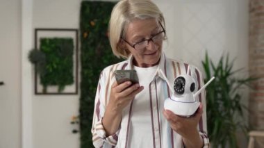 Modern güvenlik kamerası ve cep telefonunu elinde tutan yetişkin bir beyaz kadın uygulamayı çalıştırmaya çalışıyor ve internetteki ağır çekimde okuma talimatlarını ayarlıyor.