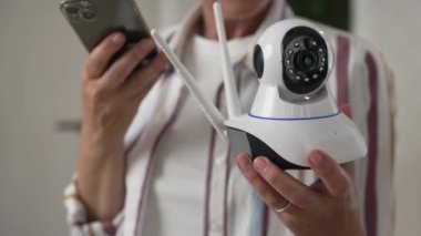 Modern güvenlik kamerası ve cep telefonunu elinde tutan yetişkin bir beyaz kadın uygulamayı çalıştırmaya çalışıyor ve internetteki ağır çekimde okuma talimatlarını ayarlıyor.