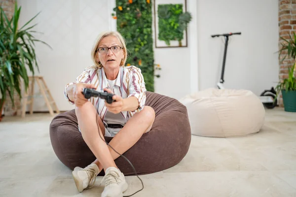 一位成熟的高加索金发女人 带着眼镜玩电子游戏 一边坐在家里 一边真正享受休闲的概念复制空间 一边笑着庆祝胜利复制空间 — 图库照片