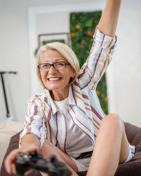 メガネプレイビデオゲームコンソールを使用してジョイスティックコントローラーを使用しながら 自宅で座っている間に成熟した白人ブロンドの女性レジャーコンセプトコピースペース幸せな笑顔祝い勝利コピースペース — ストック写真