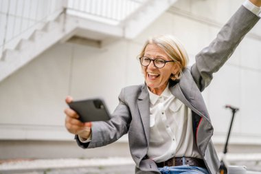 Olgun, sarışın, beyaz bir kadın açık havada mutlu bir şekilde oturup video oyunları oynuyor, cep telefonu kullanıyor, eğleniyor akıllı telefon kullanıyor, internet üzerinden fotokopi çekiyor, gözlük takıyor ve tişört giyiyor.