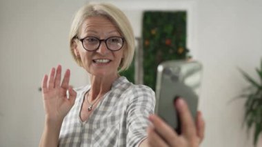 Olgun beyaz kadın akıllı cep telefonu kullanıyor. Evde, aydınlık odada ya da iş yerinde, gerçek insanlar yavaş çekim yapıyor.