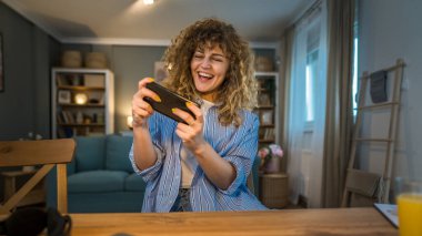 Kıvırcık saçlı yetişkin bir beyaz kadın mutlu bir şekilde evinde oturup video oyunları oynarken boş zamanlarında fren yapıyor cep telefonu kullanıyor akıllı telefon kullanıyor ve internet üzerinden eğlenceli fotokopi çekiyorlar.