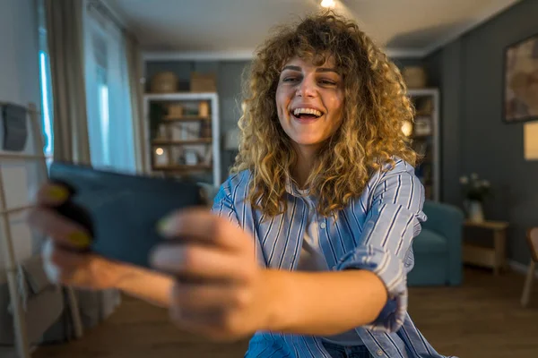 巻き毛のある大人の白人女性が自宅で幸せな笑顔でビデオゲームをプレイしながら 楽しい携帯電話の使用を持っている仕事のレジャー活動からブレーキを取るスマートフォンは楽しいオンラインコピースペースを持っています — ストック写真