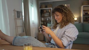 Kıvırcık saçlı yetişkin bir beyaz kadın evde mutlu bir şekilde oturup video oyunları oynarken boş zamanlarında eğleniyor cep telefonu kullanıyor, akıllı telefonları kullanıyor ve yavaş çekimde eğleniyor.