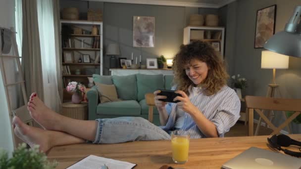 一位头发卷曲的高加索成年女子坐在家里开心地笑着玩电子游戏 同时在工作休闲活动中刹车玩得开心 — 图库视频影像