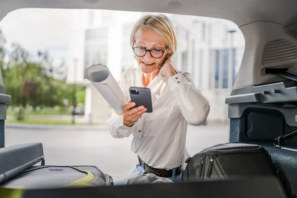 ある成熟したブロンドの美しいビジネス女性旅行コンセプトスマートフォンで女性の話は彼女の車の後ろから荷物を取りながら移動したり 目的地に到着したり 実際の人々がスペースをコピーします — ストック写真