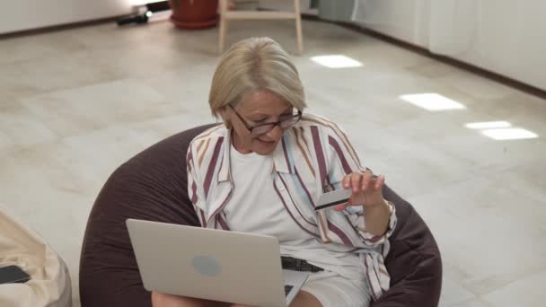 ある成熟した女性は オンラインショッピングのための家庭用クレジットカードやデビットカードを使用して座っているインターネットストア物事を購入するラップトップコンピュータを使用して実際の人々遅い動き — ストック動画