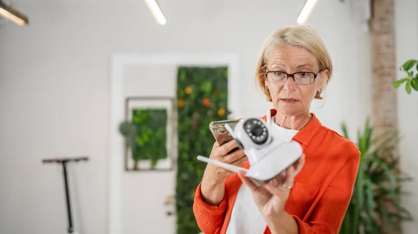 家庭の近代的なセキュリティ監視カメラとアプリのコピースペースをインストールして調整しようとする携帯電話を保持している成熟した白人女性 — ストック写真