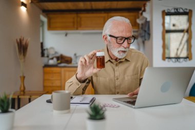 Son sınıf beyaz erkek büyükbaba evde oturup ağrı kesici veya vitamin takviyesi paketlerini tutuyor. Bilgisayardan sipariş edilen dizüstü bilgisayarın yanında fotokopi için hazır bekliyor.