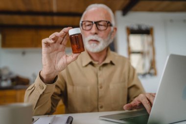 Son sınıf beyaz erkek büyükbaba evde oturup ağrı kesici veya vitamin takviyesi paketlerini tutuyor. Bilgisayardan sipariş edilen dizüstü bilgisayarın yanında fotokopi için hazır bekliyor.