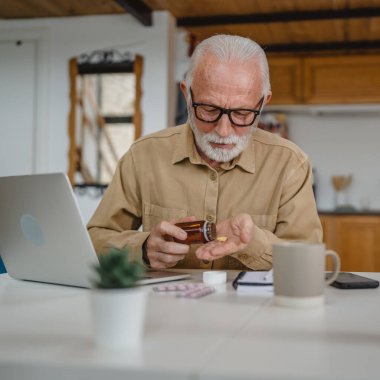 Son sınıf beyaz erkek büyükbaba evinde oturup ağrı kesici ya da vitamin takviyesi paketini tutuyor. İlaç almaya hazır.