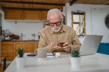 Son sınıf beyaz erkek büyükbaba evde oturup ağrı kesici veya vitamin takviyesi paketlerini tutuyor. Bilgisayardan sipariş edilen dizüstü bilgisayarın yanında fotokopi için hazır bekliyor.