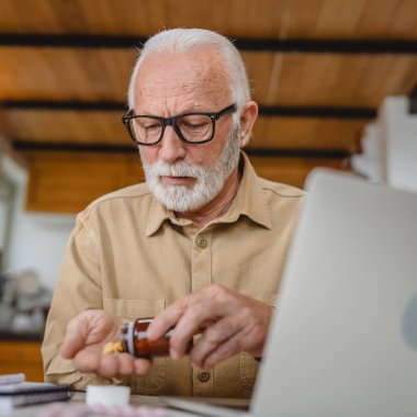 Son sınıf beyaz erkek büyükbaba evinde oturup ağrı kesici ya da vitamin takviyesi paketini tutuyor. İlaç almaya hazır.