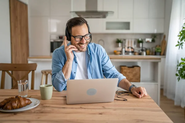 一位来自高加索的男性自由职业者在家里工作 头上戴着耳机 头戴手提电脑 顾客对他的快乐微笑充满自信 — 图库照片