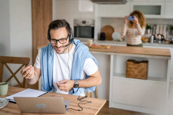 一名男子在家里工作 头上戴着耳机 头戴手提电脑 他笑得很开心 相信妻子会站在他的后面 — 图库照片