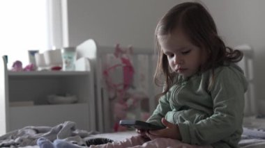 Küçük beyaz bir kız çocuğu akıllı telefonu tutarken evde ağır çekim teknolojisi ve büyüyen kavram olan gerçek insanlar yatağında oturuyor.